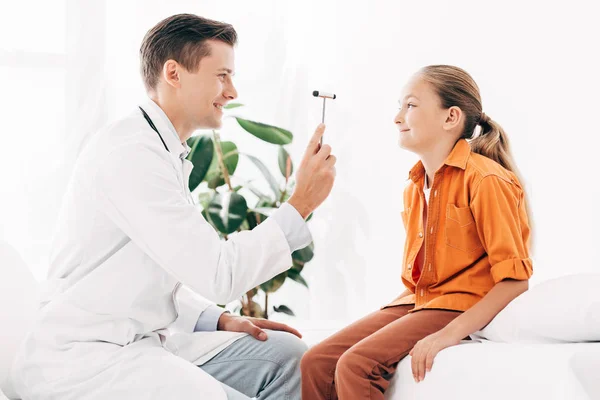 Pediatra sorridente em casaco branco examinando criança com martelo reflexo — Fotografia de Stock