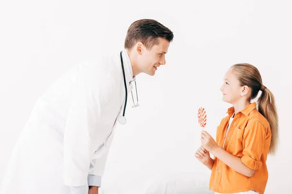 Lächelnder Kinderarzt mit Stethoskop und Kind mit Lutscher schauen einander isoliert auf weißem Grund an — Stockfoto