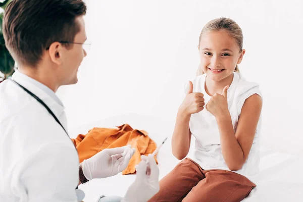 Pediatrist holding syringe and smiling child showing thumbs up — Stock Photo