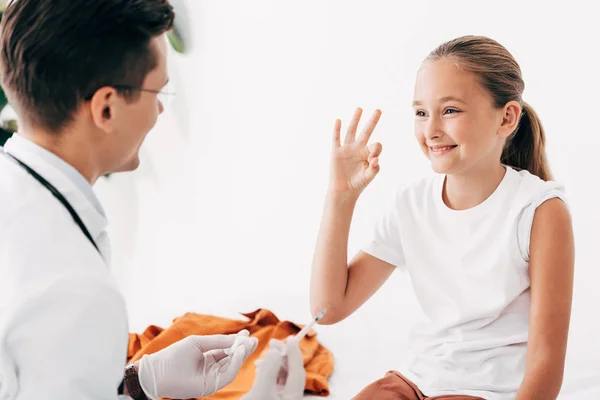 Pediatrist holding syringe and smiling child showing okay sign — Stock Photo