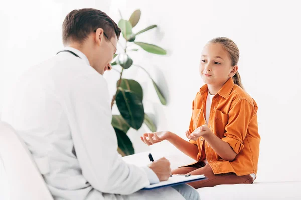 Kinderarzt schreibt in Klemmbrett, während er Kind in Klinik untersucht — Stockfoto