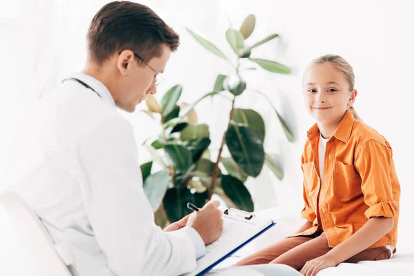 Kinderarzt schreibt in Klemmbrett, während er Kind in Klinik untersucht — Stockfoto
