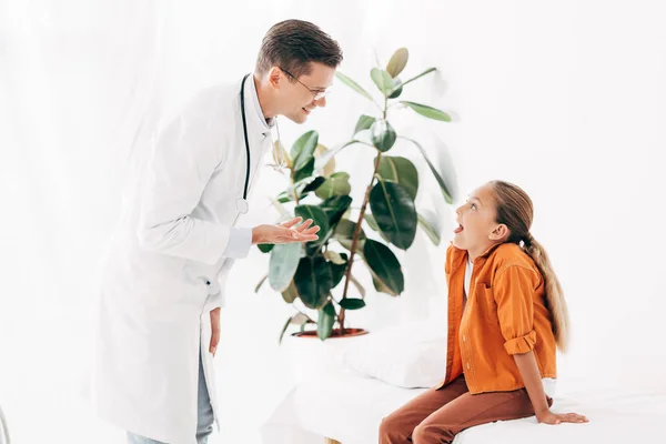 Pediatra sonriente con abrigo blanco y niño mirándose en la clínica - foto de stock