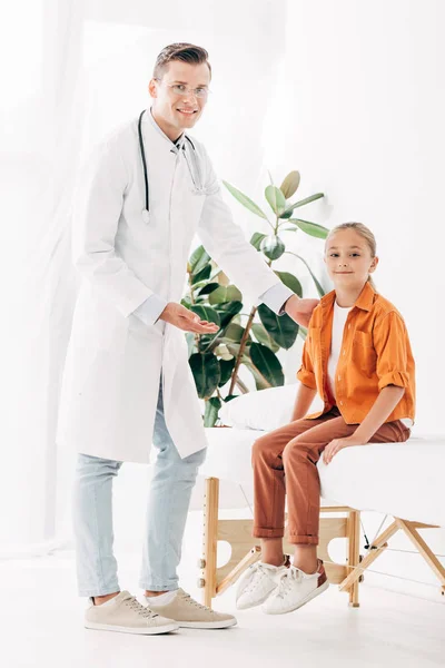 Visão de comprimento total do pediatra sorridente em casaco branco e criança na clínica — Fotografia de Stock