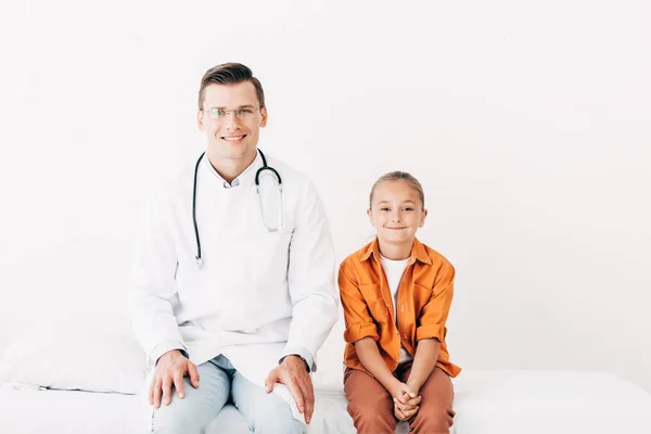 Vista frontal del pediatra sonriente con abrigo blanco y niño sentado en la cama del hospital en la clínica - foto de stock