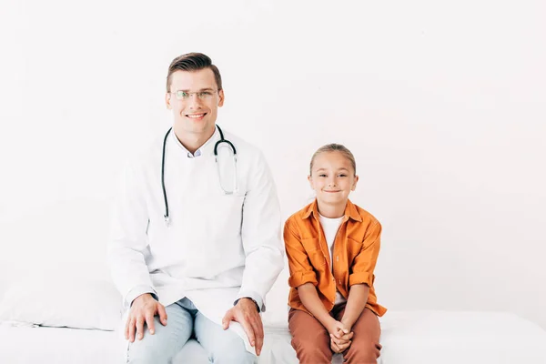 Front view of smiling pediatrist in white coat and child sitting on hospital bed in clinic — Stock Photo