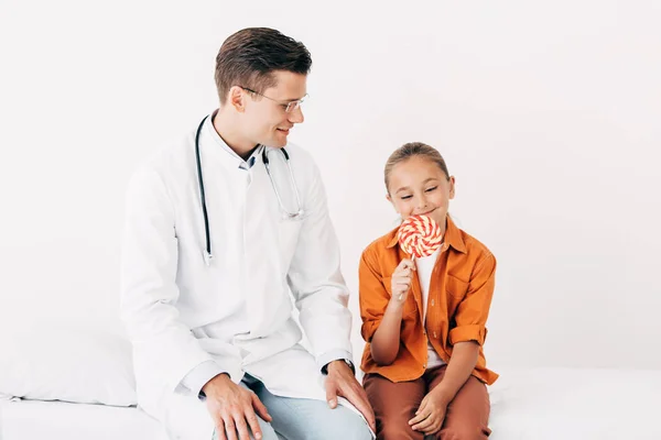 Усміхнений педіатр у білому пальто і дитина з льодяником сидить на лікарняному ліжку в клініці — стокове фото