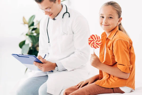 Pediatrist writing in clipboard and child holding lollipop — Stock Photo