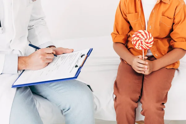 Ausgeschnittene Ansicht des Kinderarztes, der in Klemmbrett schreibt und Kind Lutscher hält — Stockfoto
