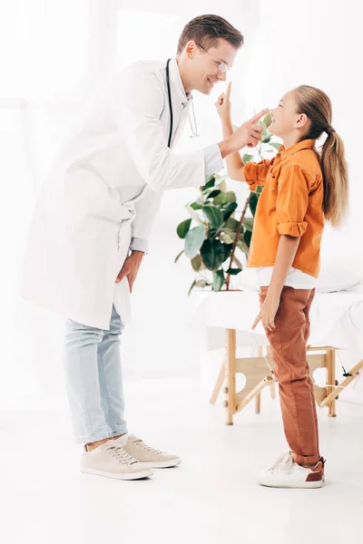 Full length view of smiling pediatrist in white coat and child in clinic — Stock Photo