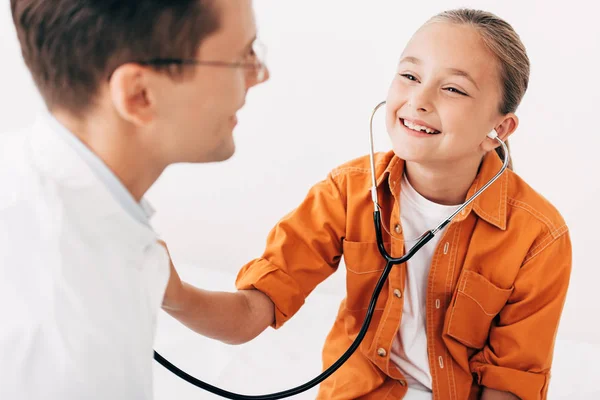 Niño sonriente examinando médico con estetoscopio aislado en blanco - foto de stock