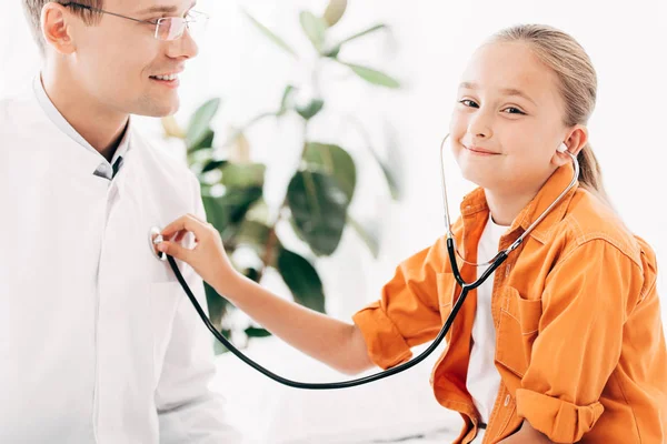 Улыбающийся ребенок осматривает врача стетоскопом в клинике — стоковое фото