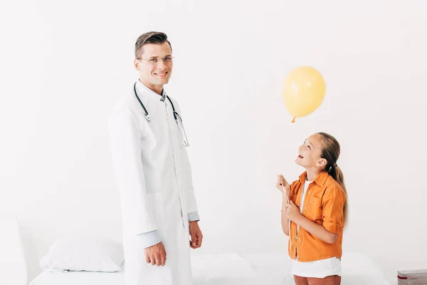 Улыбающийся педиатр в белом халате и счастливый ребенок с воздушным шаром в клинике — стоковое фото