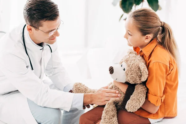 Pediatra in cappotto bianco e bambino con orsacchiotto che si guarda in clinica — Foto stock