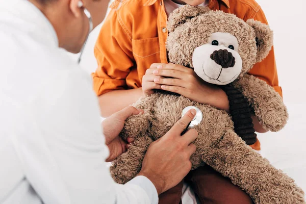 Vista parziale di bambino e pediatra in cappotto bianco esaminando orsacchiotto con stetoscopio — Foto stock