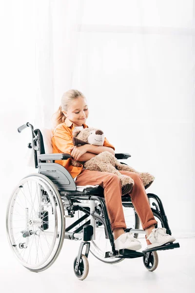 Enfant souriant tenant ours en peluche et assis sur un fauteuil roulant — Photo de stock