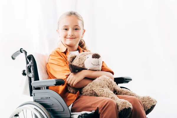 Niño sonriente sosteniendo oso de peluche y sentado en silla de ruedas - foto de stock