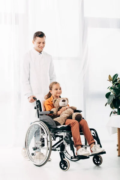 Vue pleine longueur du pédiatre en manteau blanc et enfant avec ours en peluche sur fauteuil roulant — Photo de stock