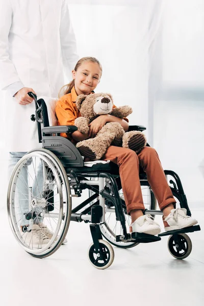 Vista parcial de pediatra en bata blanca y niño con osito de peluche en silla de ruedas - foto de stock