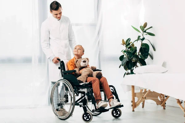 Vista completa del pediatra en abrigo blanco y niño con oso de peluche en silla de ruedas - foto de stock