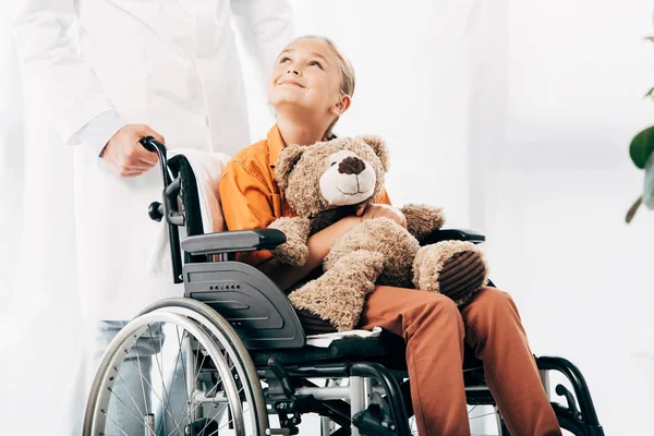 Ausgeschnittener Blick auf Kinderärztin in weißem Mantel und Kind mit Teddybär im Rollstuhl — Stockfoto