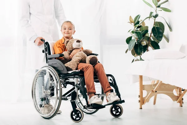 Vue recadrée du pédiatre en manteau blanc et enfant avec ours en peluche sur fauteuil roulant — Photo de stock