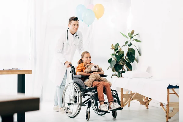 Visão comprimento total do pediatra em casaco branco e criança com ursinho de pelúcia na cadeira de rodas — Fotografia de Stock
