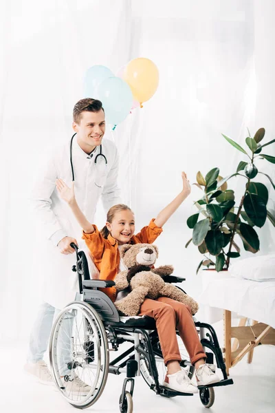 Smiling pediatrist and kid with teddy bear on wheelchair in hospital — Stock Photo