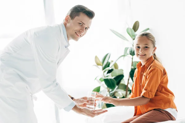 Smiling pediatrist in white coat giving glass of water to child in clinic — Stock Photo