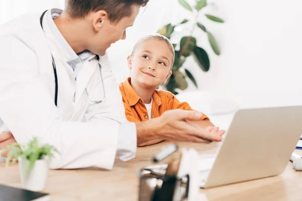 Pediatra con bata blanca y niño usando portátil en la clínica - foto de stock