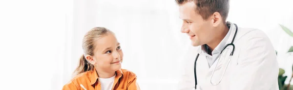Панорамний знімок усміхненої дитини та педіатра, який дивиться один на одного — стокове фото