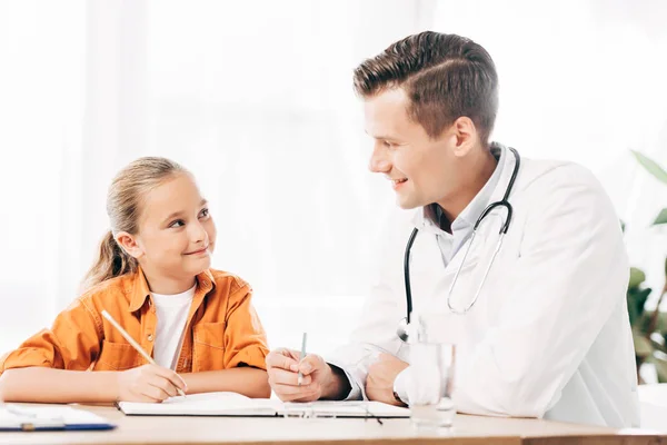Niño sonriente y pediatra mirándose mientras escriben en el cuaderno en la mesa en la clínica - foto de stock