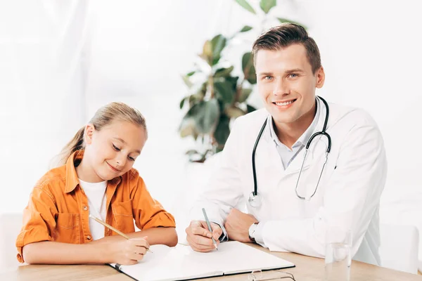 Улыбающийся ребенок и педиатр пишут в блокноте за столом в клинике — стоковое фото