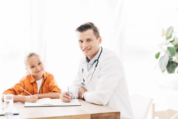 Criança sorridente e pediatra escrevem em caderno à mesa na clínica — Fotografia de Stock