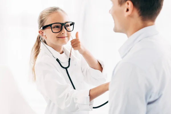 Criança em traje médico examinando paciente com estetoscópio e mostrando o polegar para cima — Fotografia de Stock