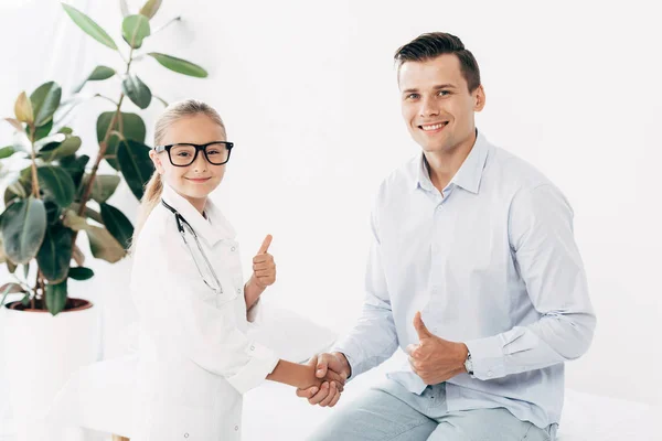 Bambino sorridente in costume medico e paziente che stringe la mano e mostra i pollici in su — Foto stock