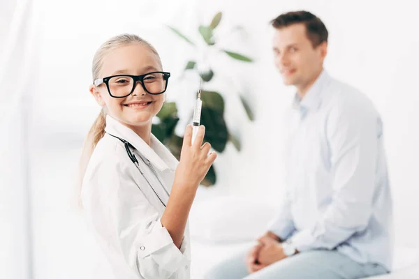 Niño sonriente con traje de médico y gafas con jeringa - foto de stock