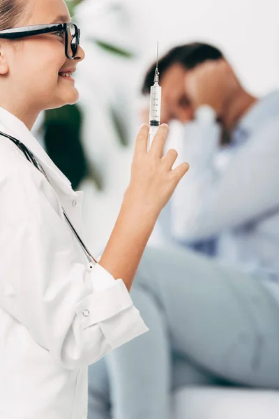 Criança sorridente em traje médico e óculos segurando seringa — Fotografia de Stock