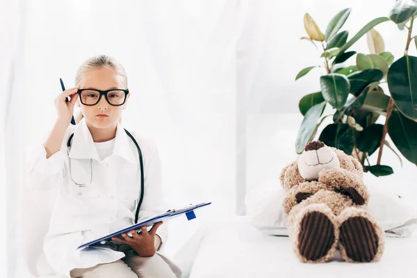Vue de face de l'enfant en costume de médecin tenant presse-papiers et stylo dans la clinique — Photo de stock