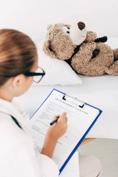 Foco seletivo da criança no traje médico examinando ursinho de pelúcia e escrita na área de transferência — Fotografia de Stock