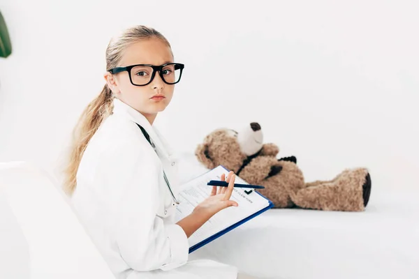 Niño en traje médico sujetando portapapeles y examinando oso de peluche - foto de stock
