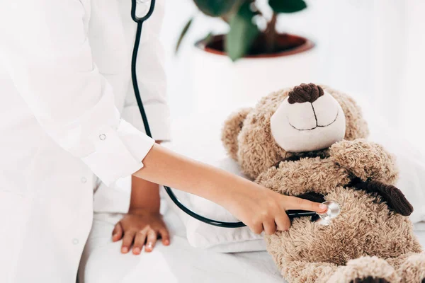 Visão parcial da criança no traje médico examinando ursinho de pelúcia com estetoscópio — Fotografia de Stock