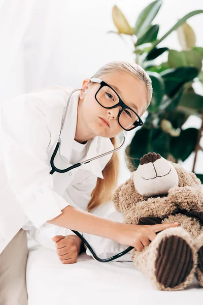 Kid in doctor costume examining teddy bear with stethoscope — Stock Photo