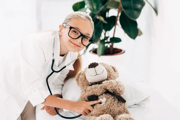 Lächelndes Kind im Arztkostüm untersucht Teddybär mit Stethoskop — Stockfoto