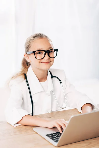 Smiling child in doctor costume using laptop in clinic — Stock Photo
