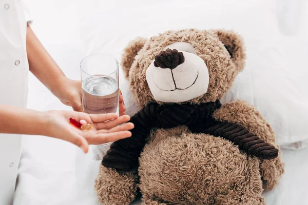Partial view of child giving glass of water and pills to teddy bear — Stock Photo