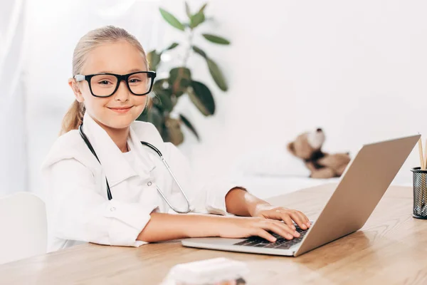 Smiling child in doctor costume using laptop in clinic — Stock Photo