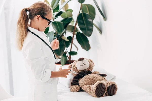 Criança em traje médico e óculos de pé perto de ursinho de pelúcia e segurando seringa — Fotografia de Stock