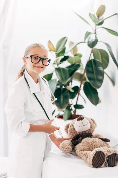 Kind in Arztkostüm und Brille steht neben Teddybär und hält Spritze — Stockfoto