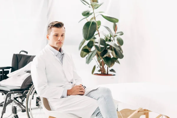 Médico pensativo de abrigo blanco sentado con las manos apretadas - foto de stock